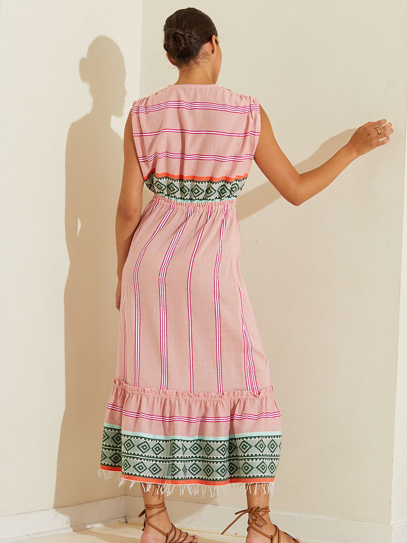 Back view of a woman standing wearing the Rosa Sleeveless Plunge Neck Dress featuring architectural details and geometric border patterns woven on a soft pink background.
