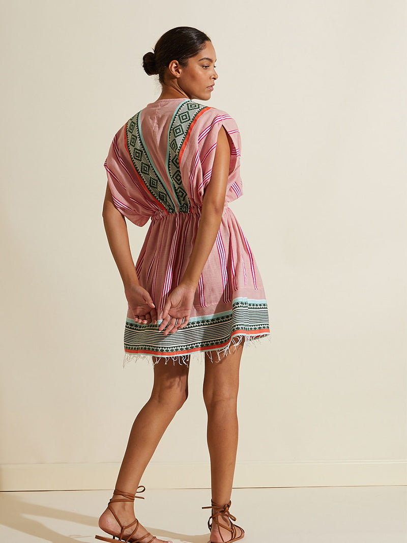 Back view of a woman wearing the Rosa Short Plunge Neck Dress featuring architectural details and geometric border patterns woven on a soft pink background.