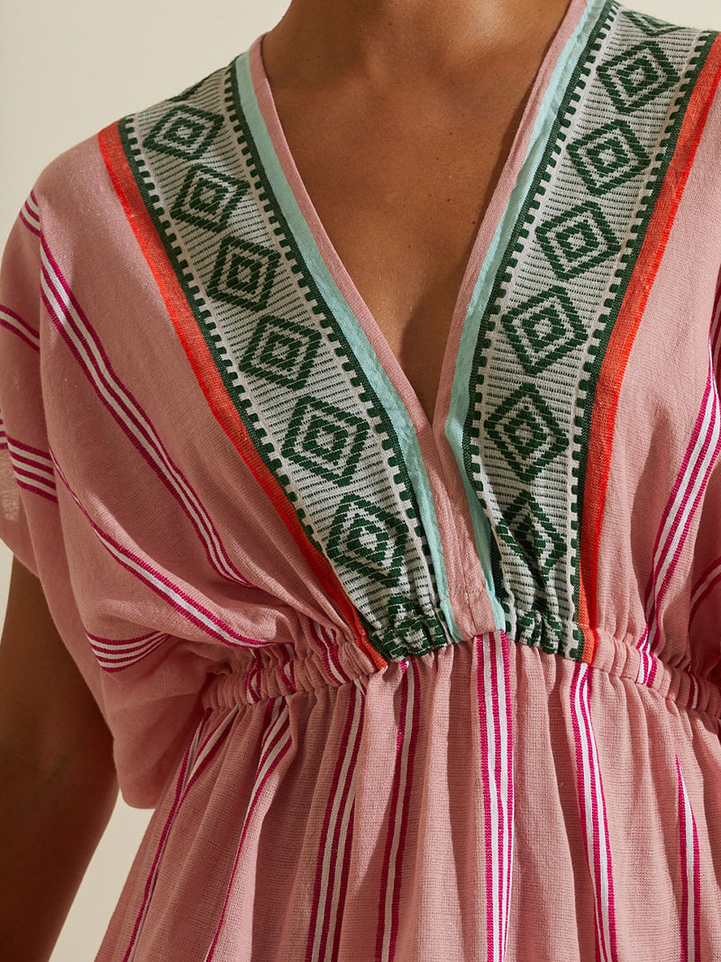 Close up on the chest of a woman wearing the Rosa Short Plunge Neck Dress featuring architectural details and geometric border patterns woven on a soft pink background.