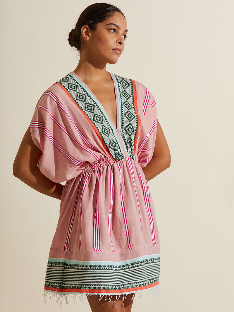 Woman standing with her hands in her back wearing the Rosa Short Plunge Neck Dress featuring architectural details and geometric border patterns woven on a soft pink background.