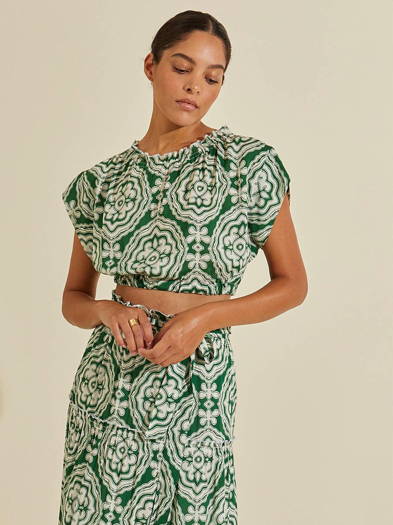 Woman standing wearing the Medallion Ruched Crop Top and matching Wrap Maxi Skirt featuring architectural white patterns on a deep green background.