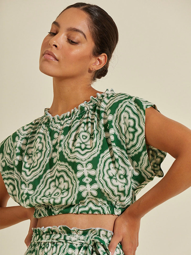 Woman standing with her hands on her hips wearing the Medallion Ruched Crop Top and matching Wrap Maxi Skirt featuring architectural white patterns on a deep green background.