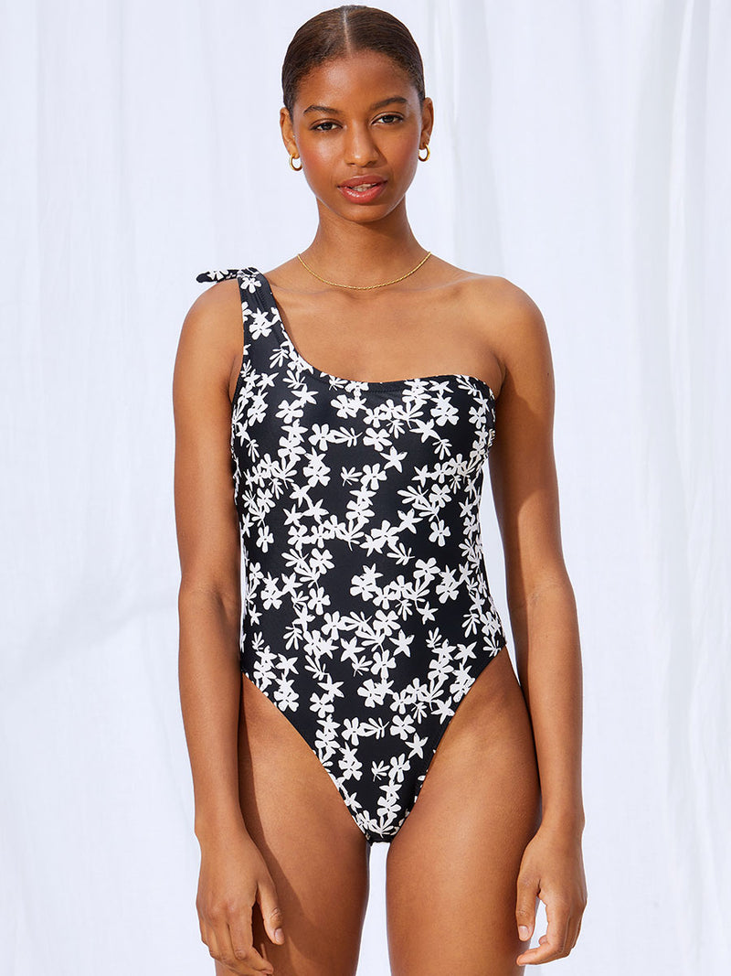 A woman standing wearing the Sea Floral One Shoulder One Piece in black featuring white allover floral print.