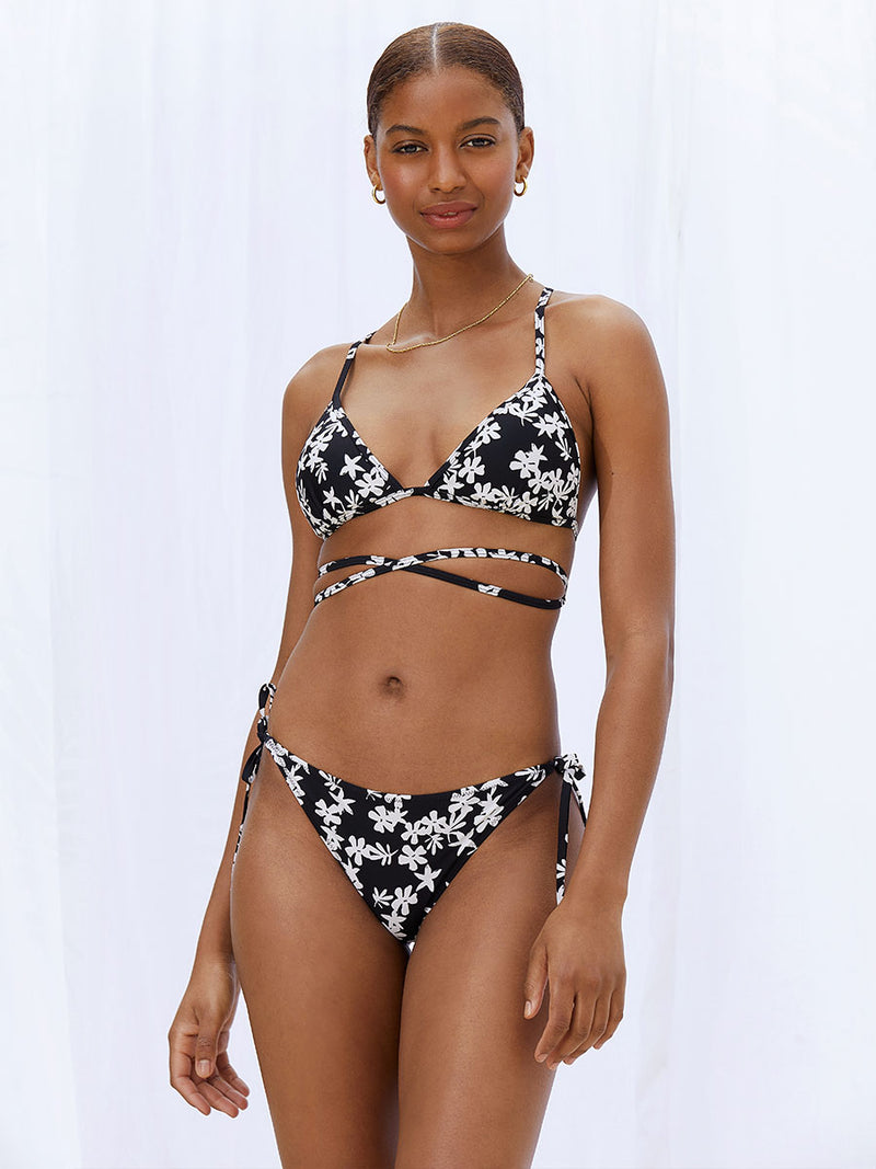 A woman standing wearing the Sea Floral Wrap Traingle Top in black featuring white allover floral print and matching string bikini bottom.