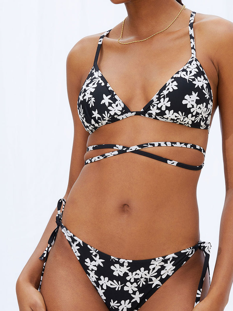 A close up view of a woman standing wearing the Sea Floral Wrap Traingle Top in black featuring white allover floral print and matching string bikini bottom.