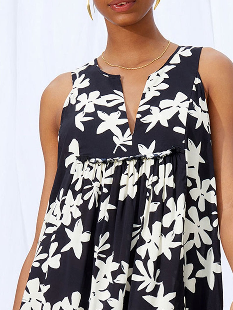 A close up view of a woman standing wearing the Sea Floral Bib Dress in Black featuring white allover floral print and a matching waist tie.