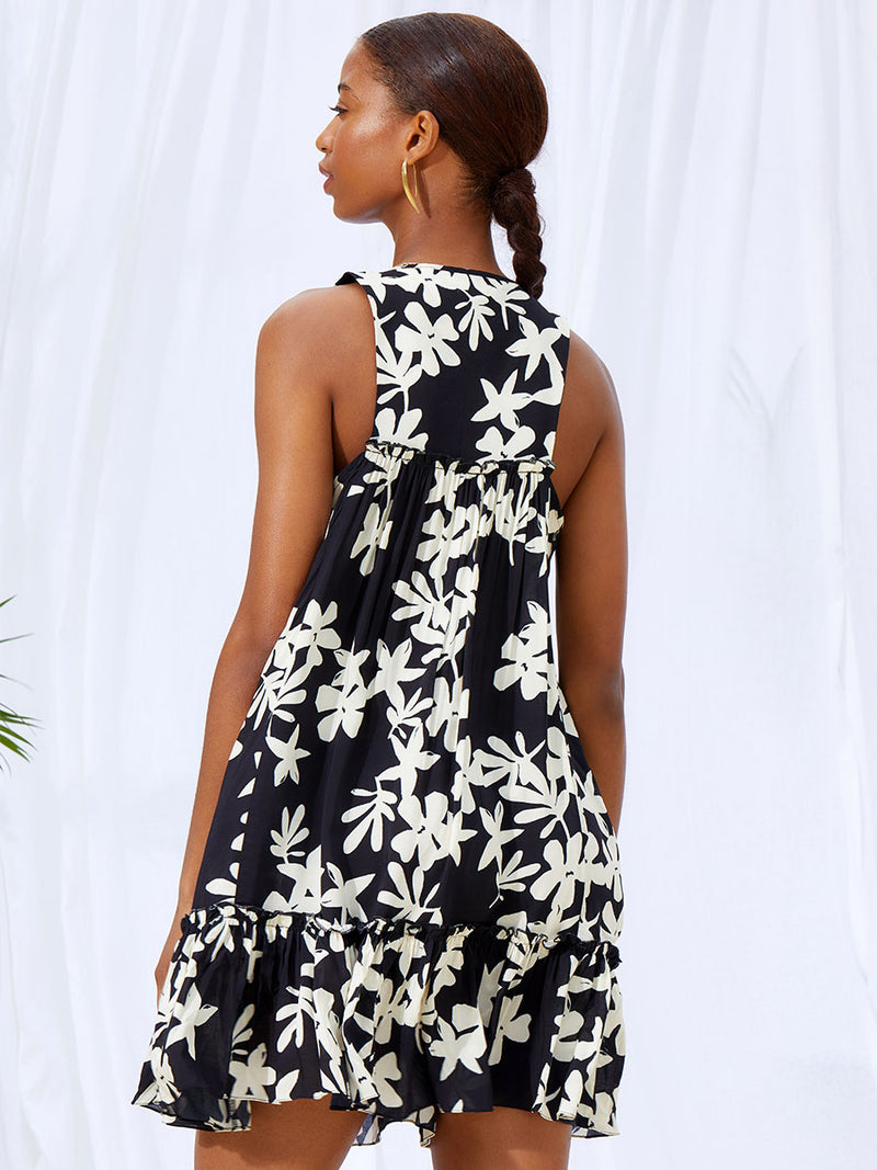 A back view of a woman standing wearing the Sea Floral Bib Dress in Black featuring white allover floral print and a matching waist tie.