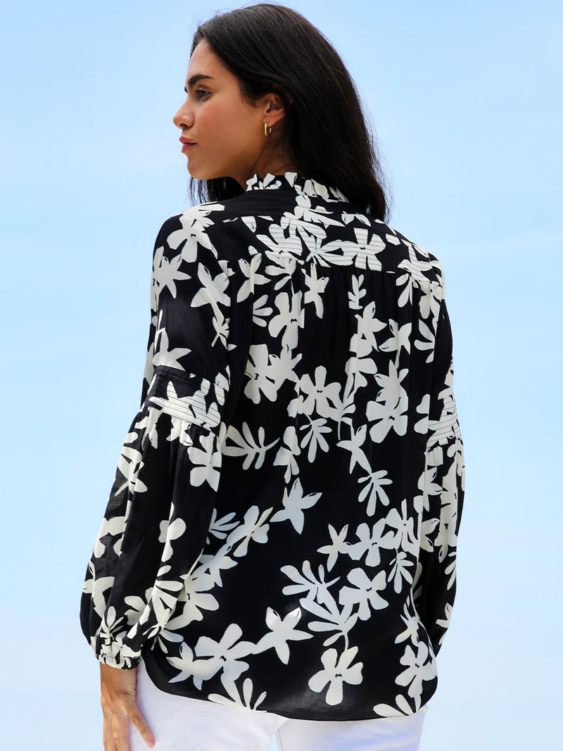 A back view of a woman standing wearing the Sea Floral Ruffle Blouse in Black featuring white allover floral print.