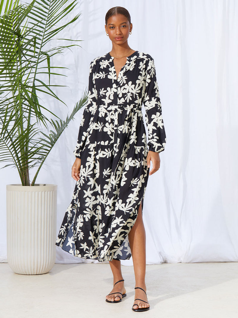 A woman standing wearing the Sea Floral Peasant Dress in Black featuring white allover floral print and a matching waist tie.