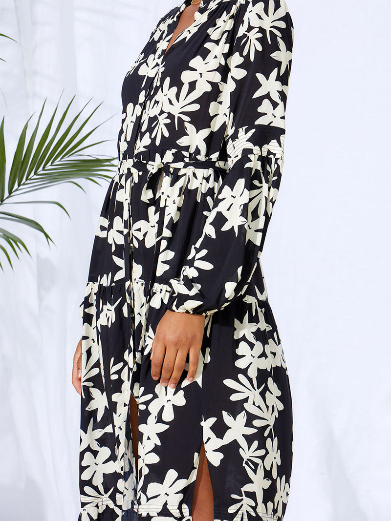 A close up side view of a woman standing wearing the Sea Floral Peasant Dress in Black featuring white allover floral print and a matching waist tie.