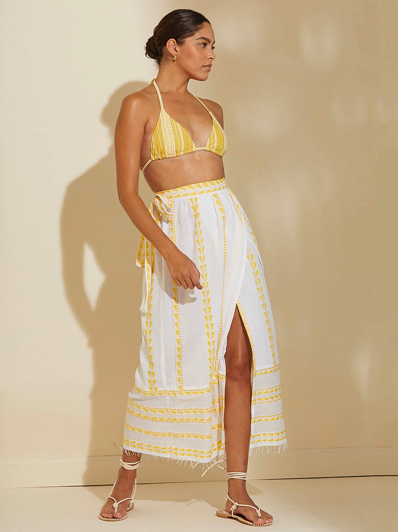 Woman standing wearing the Abeba Wrap Skirt featuring the yellow signature Tibeb pattern and gold lurex highlights on a white background and wearing the matching Luchia Triangle Top in yellow and enhanced with ivory and strands of golden shimmering lurex. 