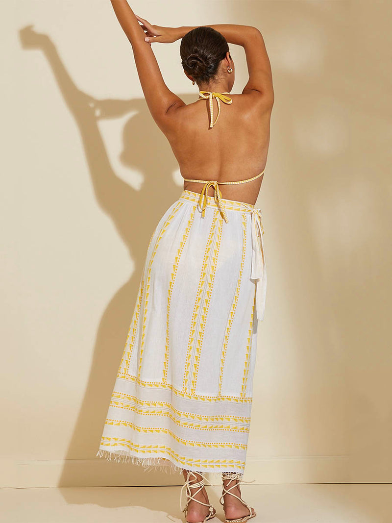 Back view of a woman standing with her arms up in the airwearing the Abeba Wrap Skirt featuring the yellow signature Tibeb pattern and gold lurex highlights on a white background and wearing the matching Luchia Triangle Top in yellow and enhanced with ivory and strands of golden shimmering lurex.