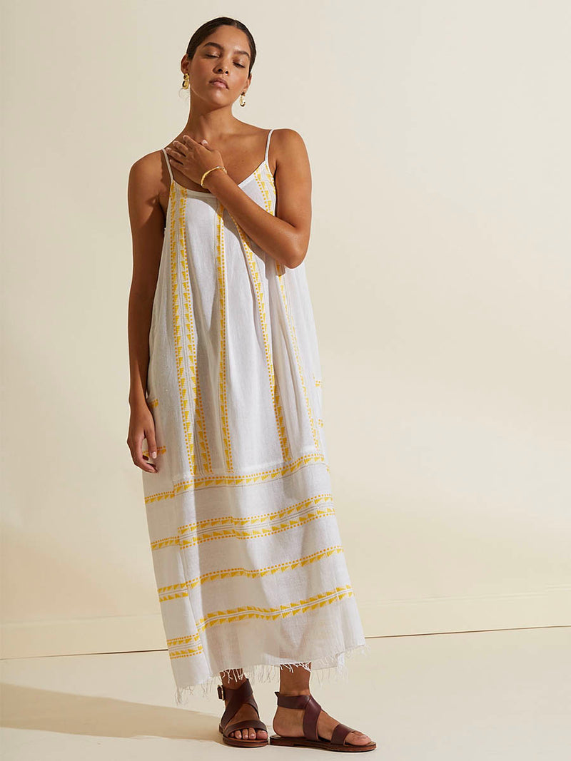 Woman standing with her hand on her neck  wearing the Abeba Slip Dress featuring the yellow signature Tibeb pattern and gold lurex highlights on a white background.