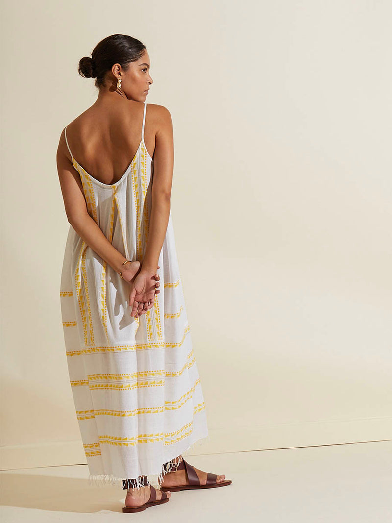 Back view of a woman standing with her hands in her back wearing the Abeba Slip Dress featuring the yellow signature Tibeb pattern and gold lurex highlights on a white background.