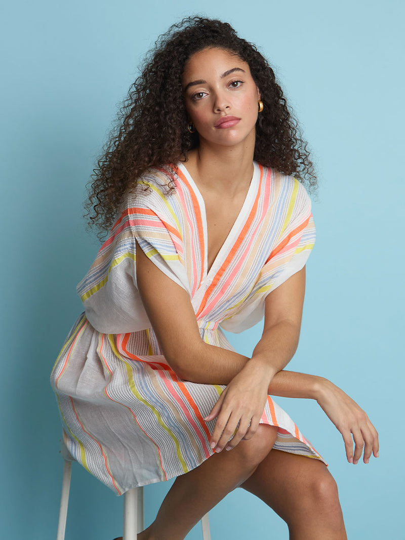 A woman sitting on a stool wearing the Tirunesh Short Plunge Neck Dress featuring pale pink, blue, yellow, orange and nude stripes