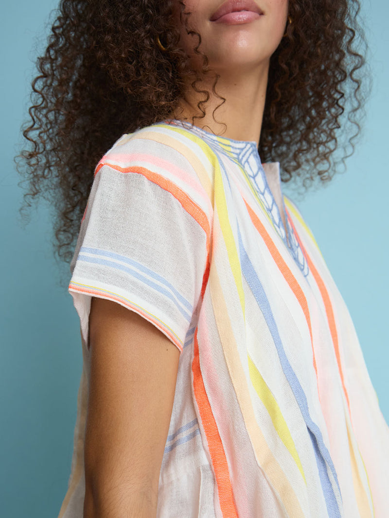 A close up view of a woman standing wearing the Tirunesh Smock Dress featuring pale pink, blue, yellow, orange and nude stripes.
