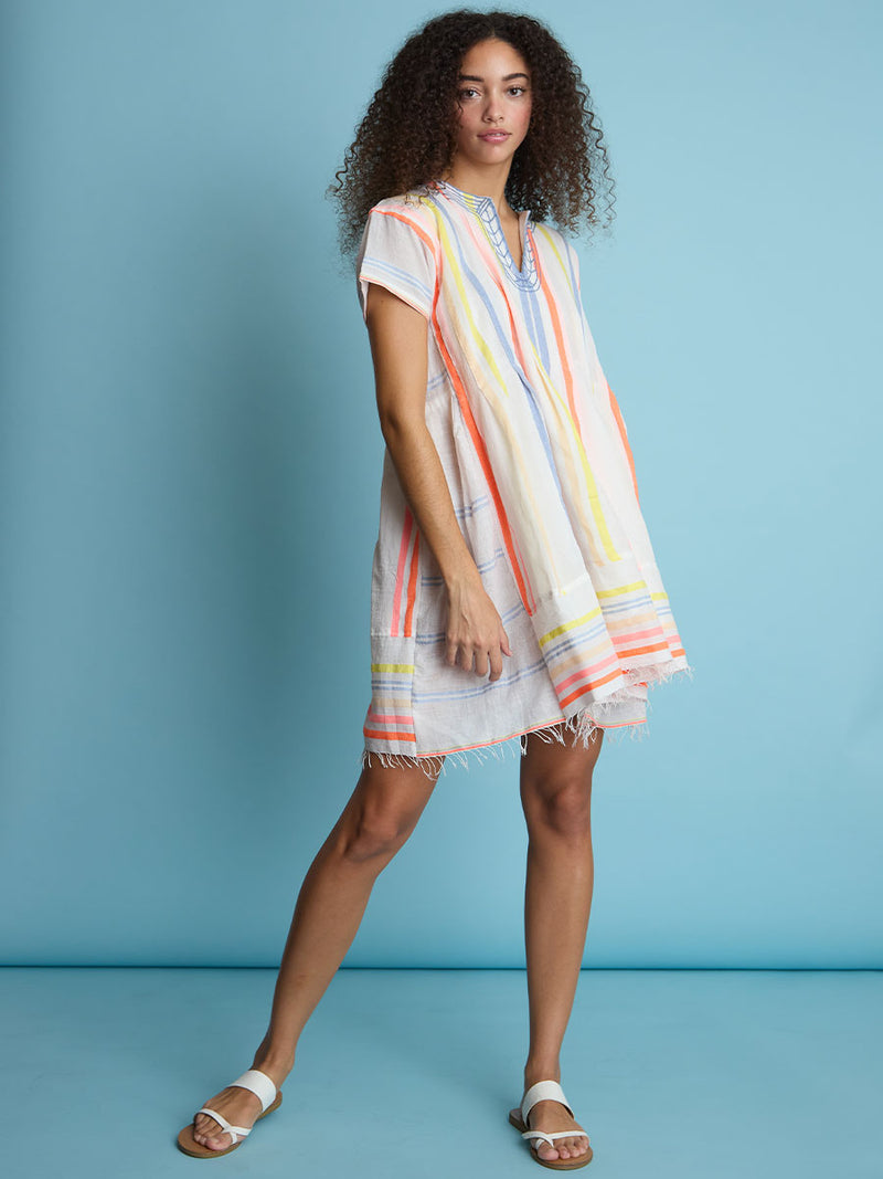 A woman standing wearing the Tirunesh Smock Dress featuring pale pink, blue, yellow, orange and nude stripes.