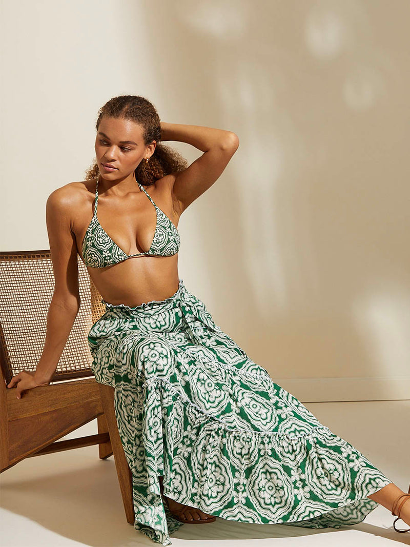 Woman sitting on a chair wearing the Medallion Maxi Skirt and Triangle Bikini Top featuring architectural white patterns on a deep green background.