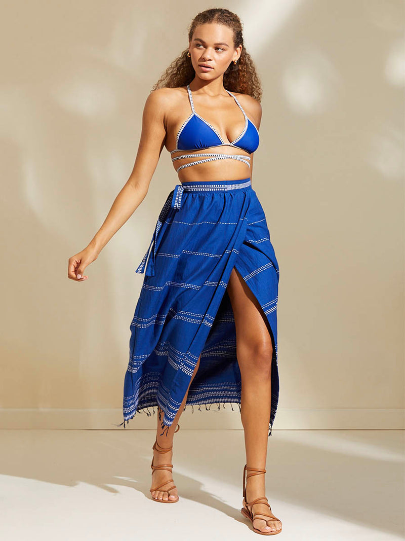 Woman walking wearing the Inku Wrap Skirt featuring textured white dots on deep blue background and matching Lena Wrap Triangle Top in bright neon blue adorned with navy and white diamond jacquard pattern on the trims